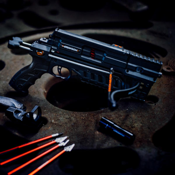 Eine schwarze AR-6 Stinger II Compact liegt ungespannt auf einem rostigen runden Eisenteil. Im Vordergrund liegen außerdem einige rote Jagdpfeile, ein Rotpunktvisier und die Steambow Taschenlampe.